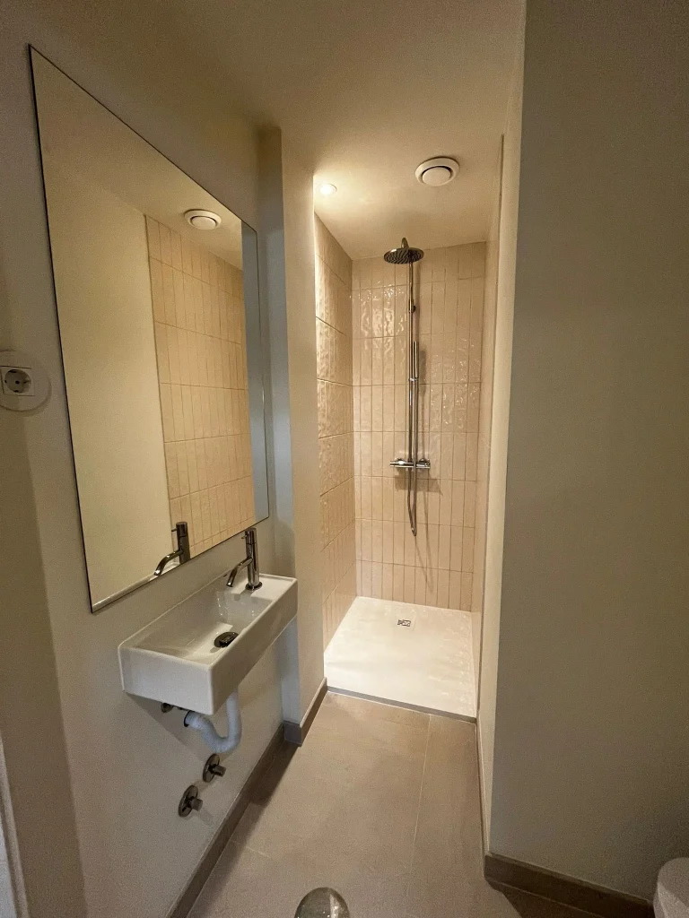Imagem de uma casa de banho pequena completamente remodelada, com design moderno e funcional. Apresenta zona de duche e lavatório branco e acabamento elegante.