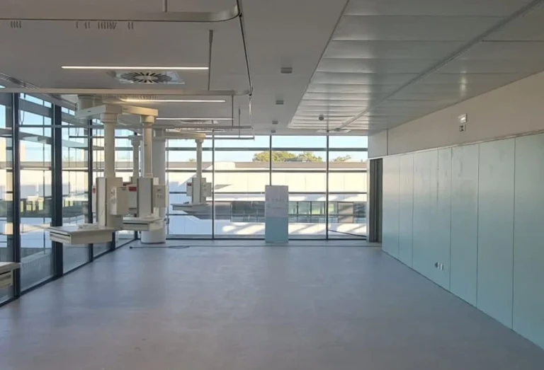 Sala clínica ampla com janelas de grande dimensão, braços técnicos suspensos no teto para suporte de equipamentos médicos e ambiente minimalista e esterilizado, preparado para procedimentos ou cuidados especializados.