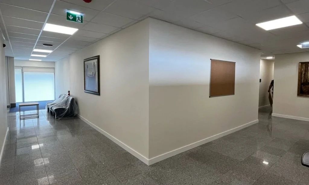 Corredor interior com pavimento cinzento polido, paredes brancas e iluminação embutida; inclui uma zona de espera com sofá protegido por plástico, mesa pequena e sinalética de emergência, transmitindo ambiente limpo e organizado.