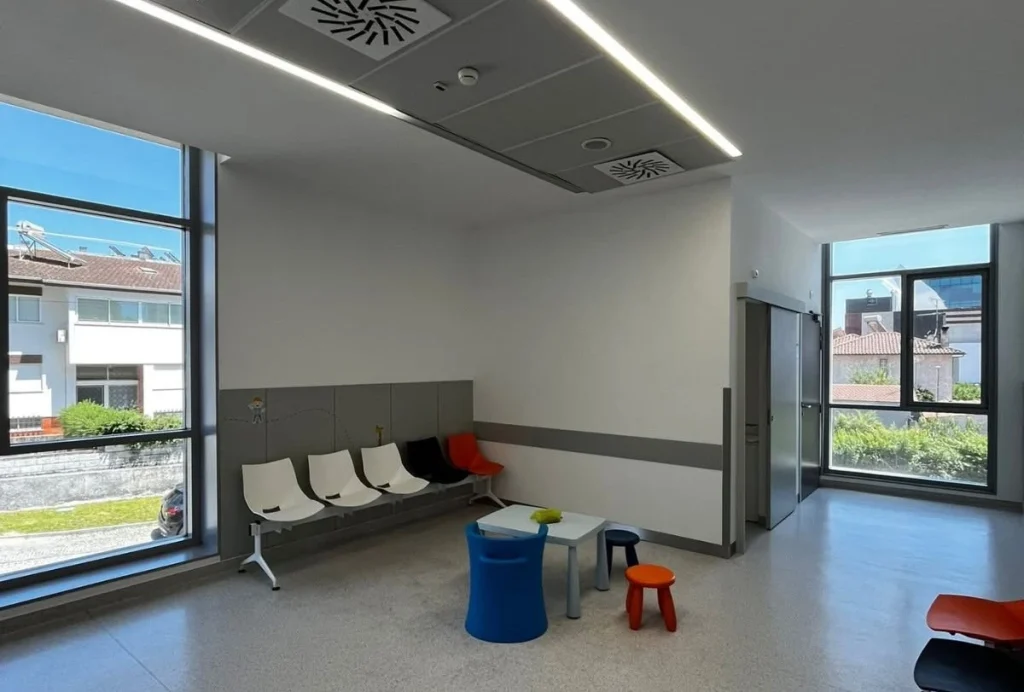 Sala de espera moderna com grandes janelas, cadeiras brancas e coloridas alinhadas e uma pequena área infantil com mesa e bancos; espaço luminoso, acolhedor e pensado para diferentes tipos de utentes.
