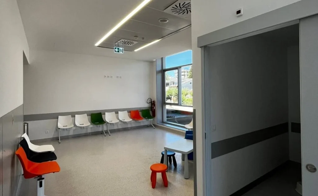 Sala de espera moderna com cadeiras coloridas alinhadas, cadeiras empilhadas e uma mesa infantil com bancos; espaço iluminado por luz natural, com extintor visível e sinalética de emergência, pensado para acolher adultos e crianças.