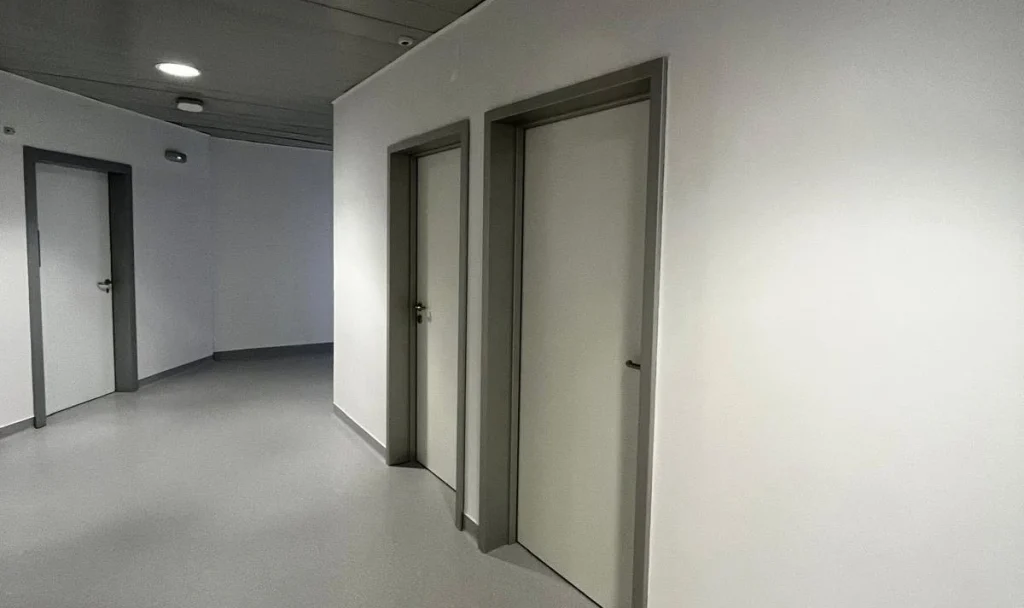 Corredor interior minimalista com pavimento cinzento, paredes brancas e três portas de acesso a gabinetes; iluminação embutida e ambiente clínico organizado e funcional.
