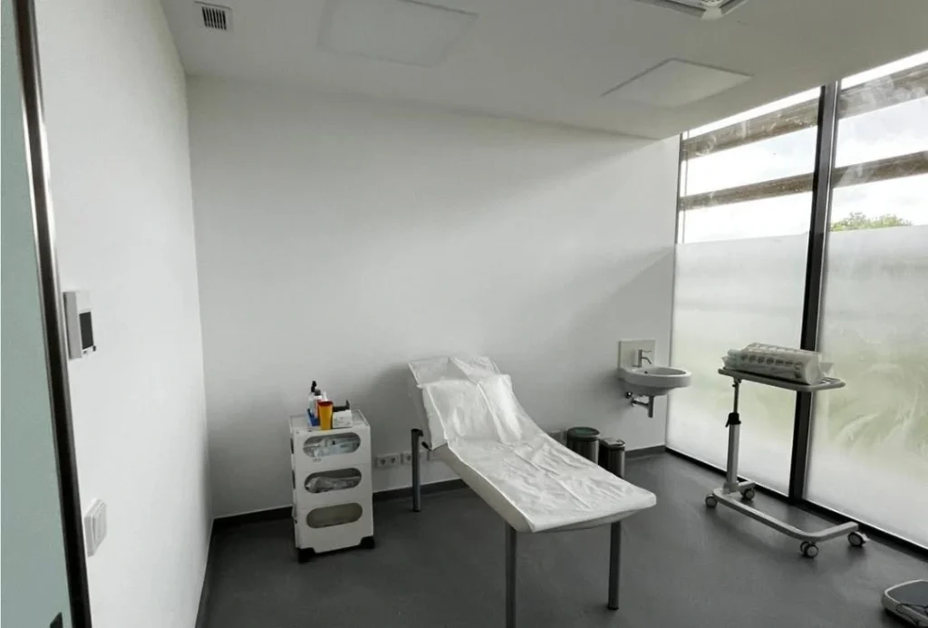 Sala de exame com marquesa coberta por proteção descartável, carrinho com consumíveis clínicos, zona de lavagem com lavatório e equipamentos de diagnóstico; ambiente esterilizado e preparado para procedimentos.